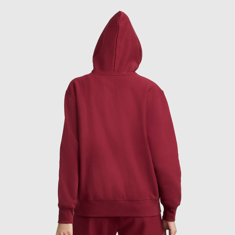 A person is shown from the back, wearing a red Champion Essentials Reverse Weave Hoodie Sweatshirt with the hood up and an embroidered C patch, set against a plain light gray background.