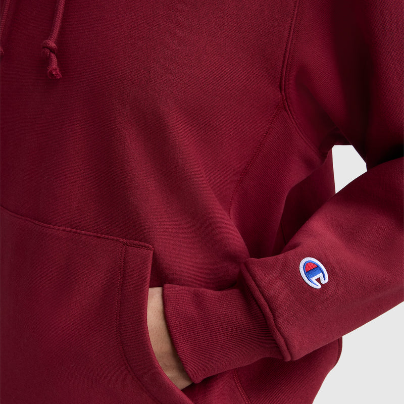 A person wearing a maroon Champion Essentials Reverse Weave Hoodie Sweatshirt with one hand in the front pocket; the sleeve displays a small embroidered C patch near the wrist.
