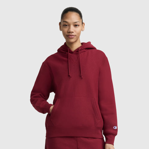 A person wearing a maroon Champion Essentials Reverse Weave Hoodie Sweatshirt with drawstrings, front pocket, and embroidered C patch stands against a plain light gray background with one hand in the pocket and a neutral expression.