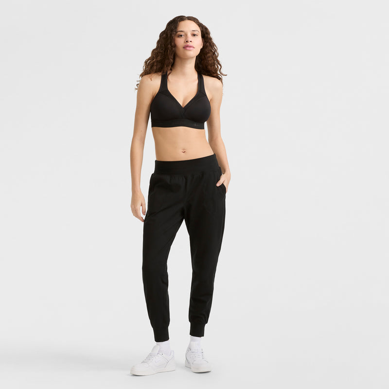 A woman with long curly hair wears the Champion Curvy Sports Bra with C Logo, black jogger pants, and white sneakers, standing against a plain white background.