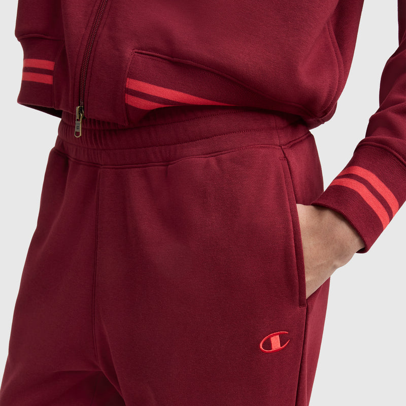A person wears Champion Pitcher Pant, C Logo joggers—maroon French terry with red striped cuffs and the Champion logo on the left pocket, standing with one hand in their elastic waistband pocket.