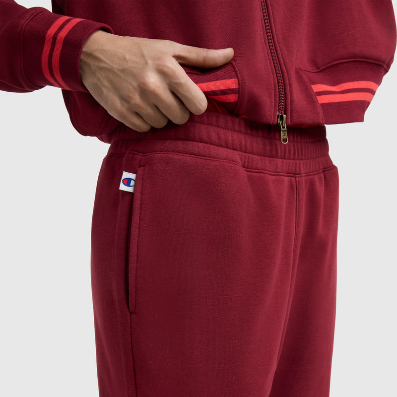 A person adjusts their jacket while wearing Champion Pitcher Pants featuring a small C logo near the pocket, striped cuffs, athletic jogger fit, and an elastic waistband.