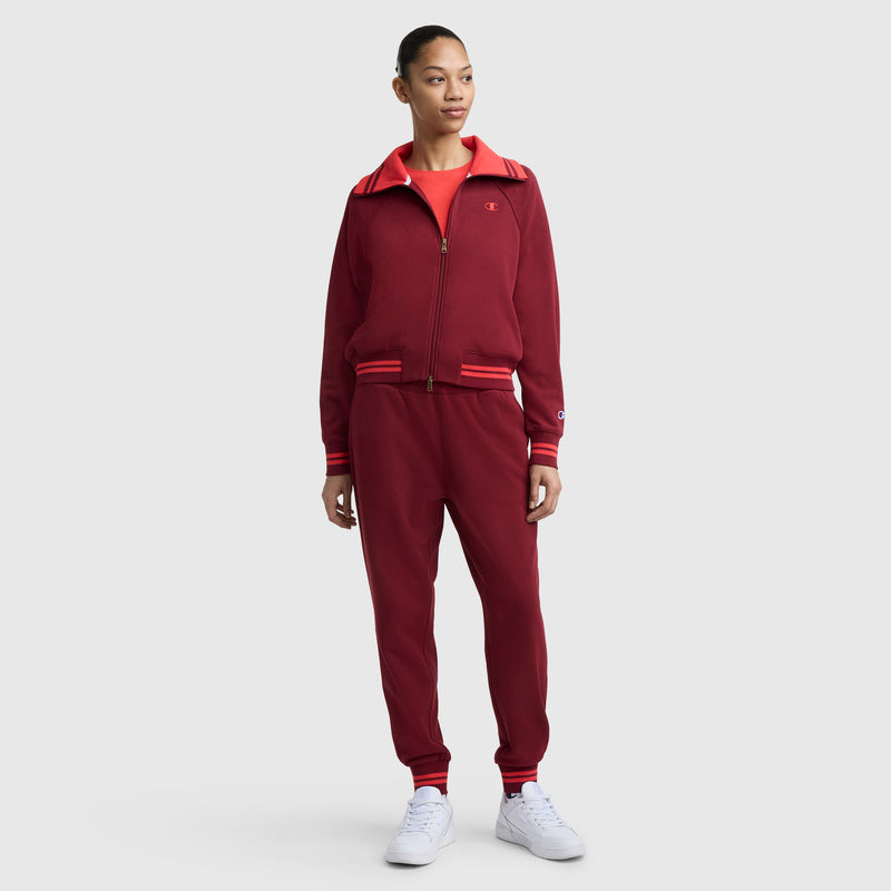 A woman stands facing forward in Champion’s Pitcher Pant with C Logo—a dark red jogger featuring an elastic waistband and red and white striped cuffs—paired with a matching zip-up jacket, red shirt, and white sneakers against a light background.