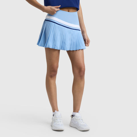 A person in the Champion Country Club Pleated Skort C Logo, paired with a blue top and white sneakers, stands against a plain light background.