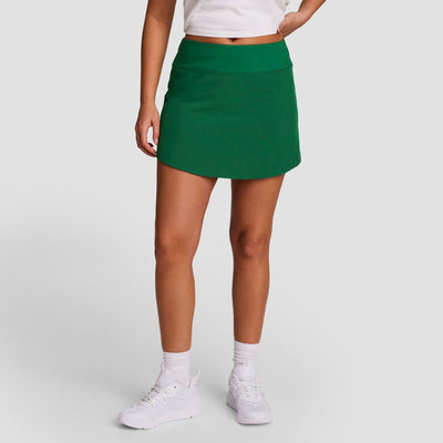 A person wears the Champion Courtside Skort, C Logo in green, paired with a white cropped top, white socks, and white sneakers, standing against a plain white background with their head out of frame.