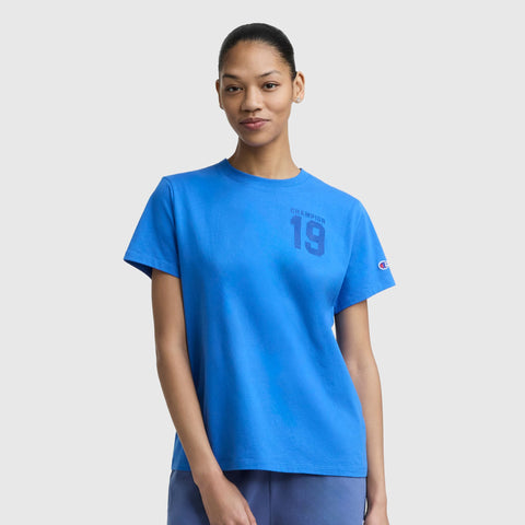 A woman in a bright blue Champion Grid Iron '19 Graphic T-Shirt with the C logo and matching pants stands against a light gray background, smiling slightly and showcasing a sporty look.