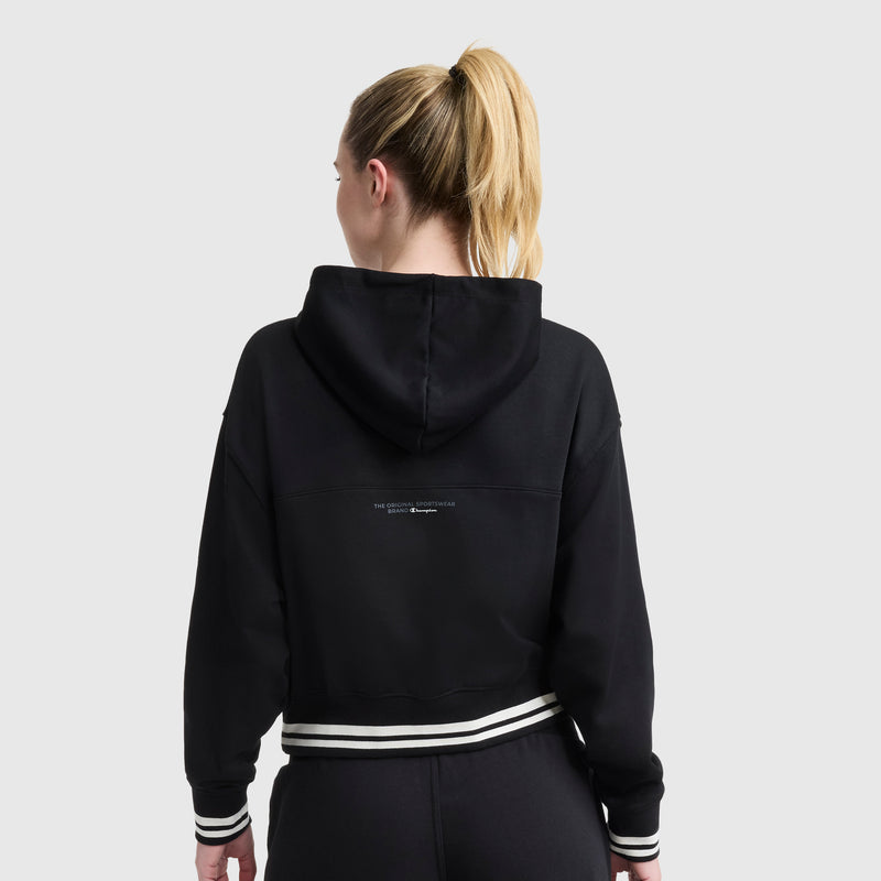 A woman with blonde hair in a ponytail models the Champion Tackle Lace Front Hoodie with a tonal embroidered C logo, paired with black pants. She stands with her back to the camera against a plain light gray background.