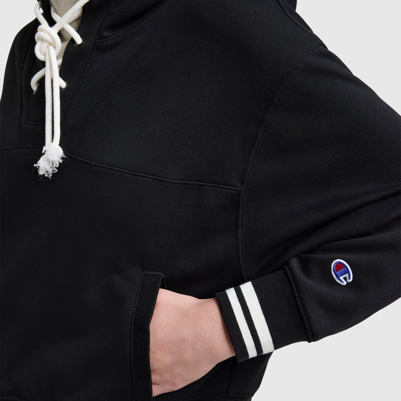 A person wears the Champion Tackle Lace Front Hoodie with a black cropped silhouette, white drawstrings, striped cuffs, a tonal embroidered C logo on the sleeve, and their hand tucked in the pocket against a plain background.