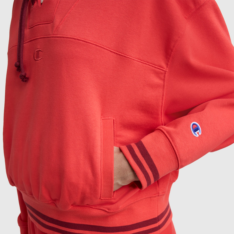 A person wears the Champion Tackle Lace Front Hoodie, featuring a bright red hue, striped cuffs and waistband, front pocket, and tonal embroidered C logo on the sleeve and chest.