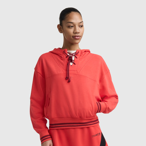 A person models the Champion Tackle Lace Front Hoodie with a tonal embroidered C logo, featuring striped cuffs and a cropped fit, standing against a plain light background with one hand in their pocket.