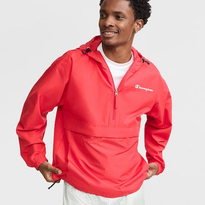 A man wearing a bright red Champion pullover windbreaker with a front zipper and a hood, standing and smiling with his hands resting in the front pocket. He is also wearing a white shirt underneath.