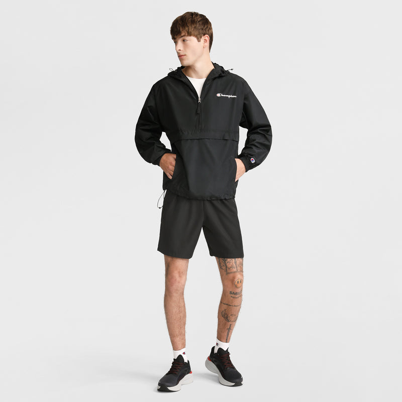 A young man in a black Champion Stadium Packable Jacket with Script Logo, black shorts, white socks, and black sneakers stands against a white background. He has visible leg tattoos and looks to the side with hands in his jacket pockets.