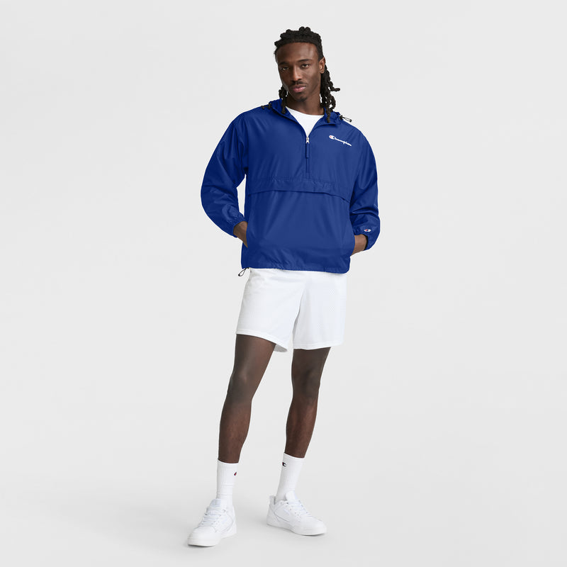 A man with long hair stands against a white background wearing a blue Champion Stadium Packable Jacket with Script Logo, white athletic shorts, socks, and sneakers. His hands are in the jacket pockets.