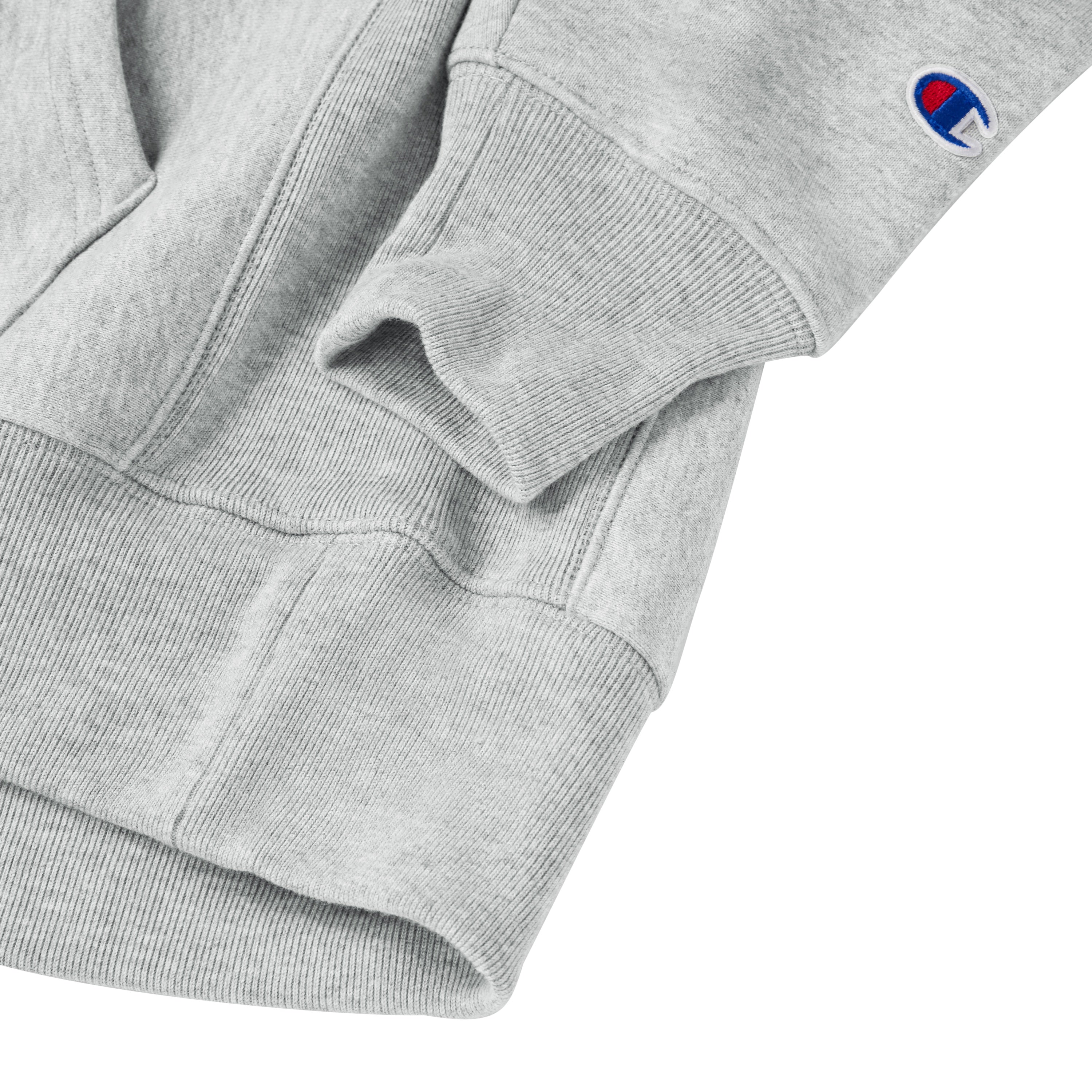 Close-up of the cuff and lower hem of a light gray sweatshirt, featuring a small embroidered Champion logo on the sleeve.