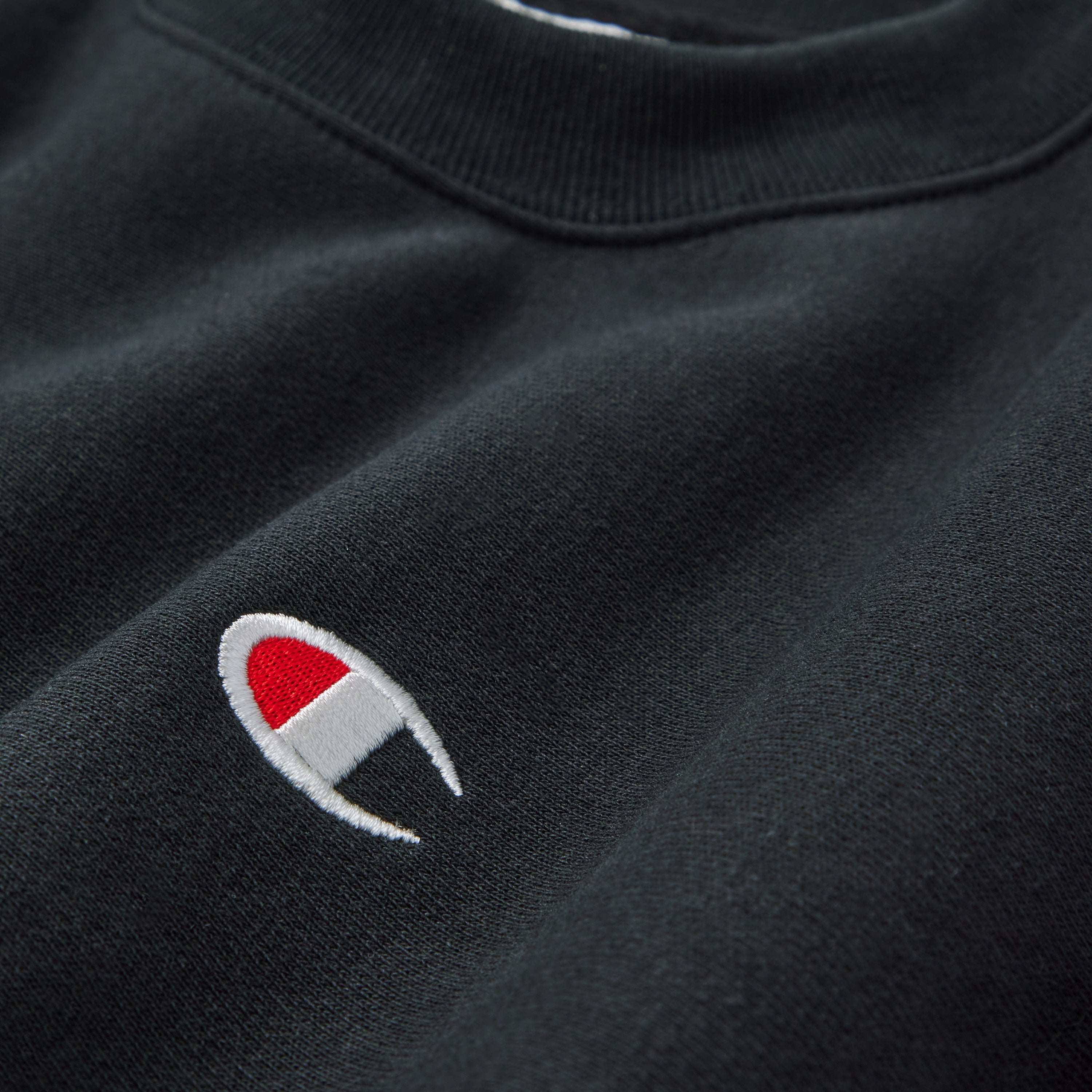 A close-up of a black sweatshirt featuring the Champion logo, which consists of a white and red C emblem, embroidered on the fabric near the chest area.