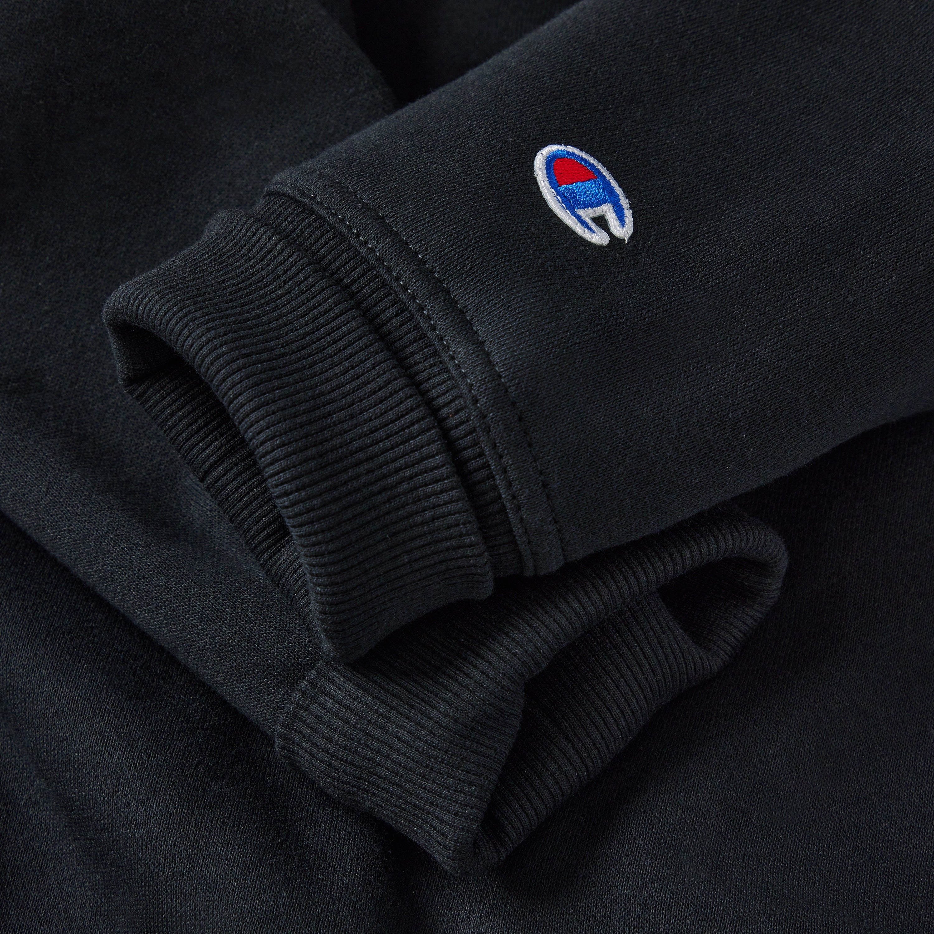 A close-up of a black sweatshirt sleeve with ribbed cuffs, featuring the Champion brand logo, which is a small embroidered C in blue, red, and white.