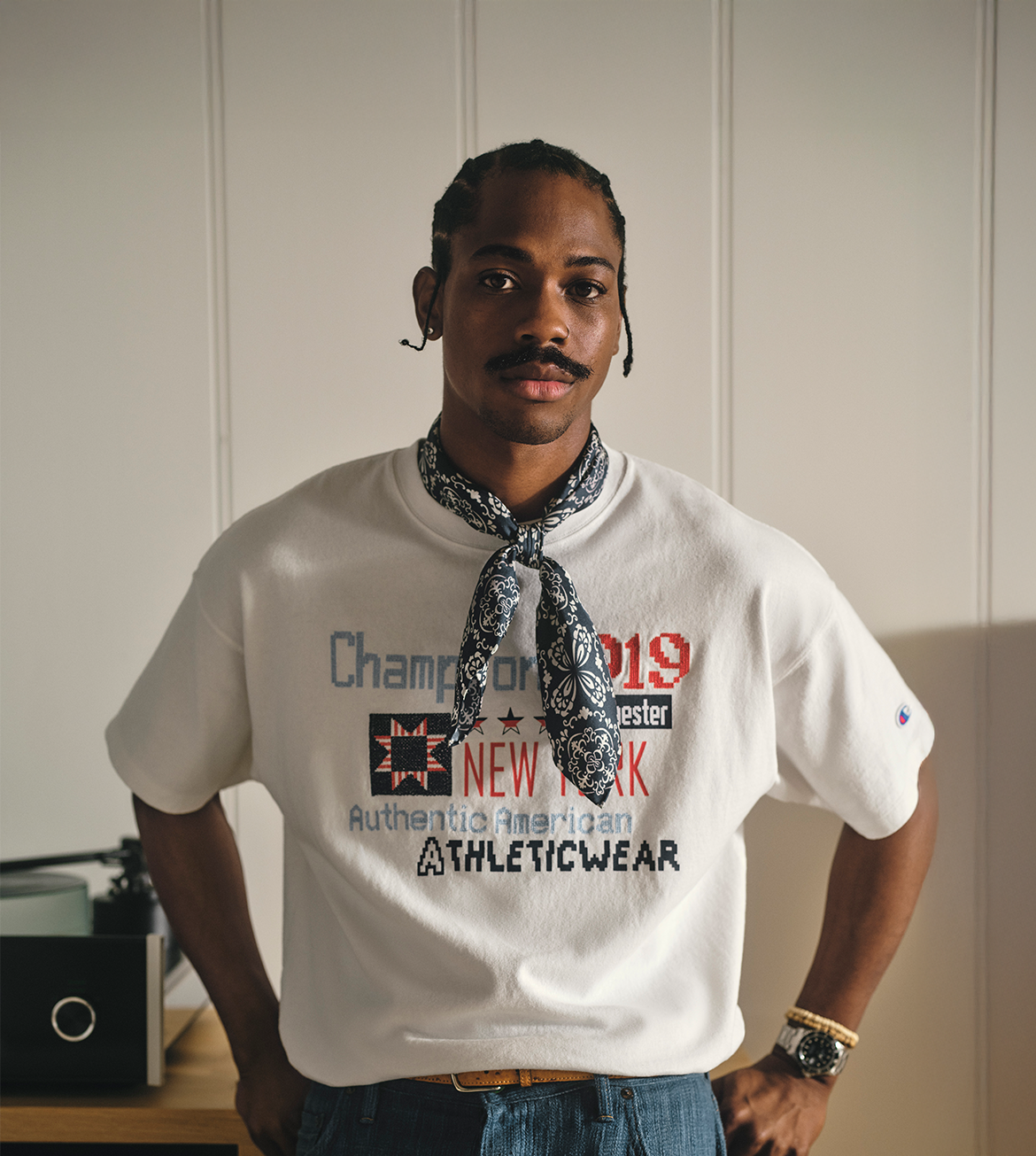 Man wearing a white Champion t-shirt with text and graphics, standing against a plain wall.