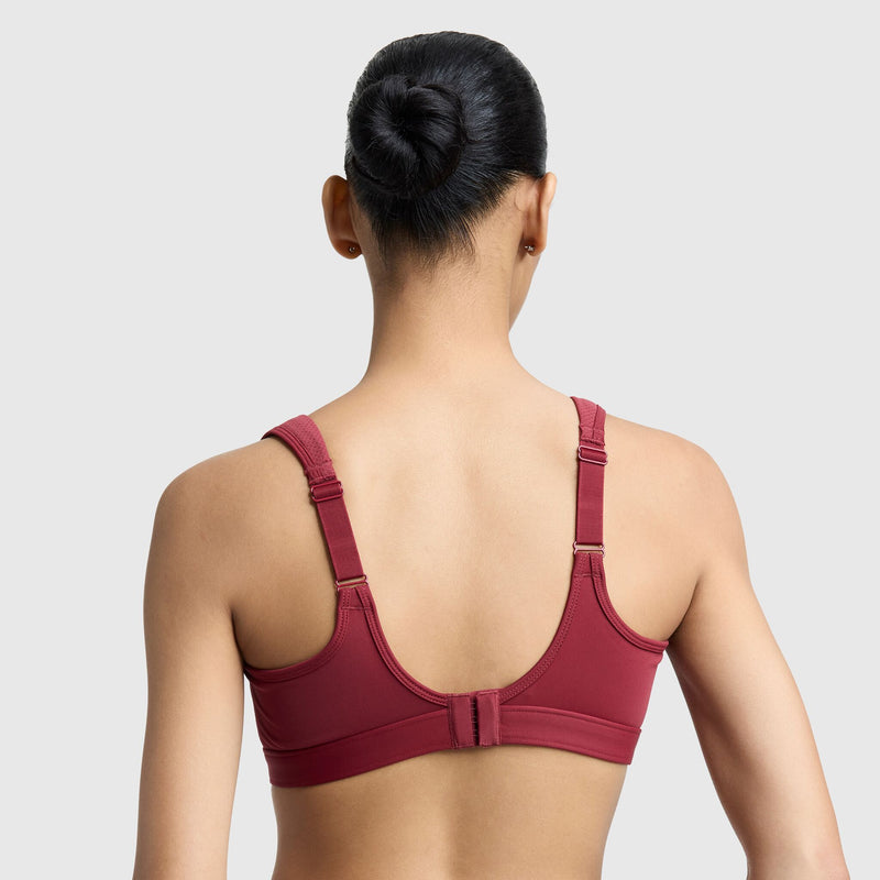 A woman with dark hair in a bun, seen from behind, wears the Champion Spot Comfort High-Impact Sports Bra, C Logo in red with adjustable straps and a hook-and-eye closure against a plain light gray background.