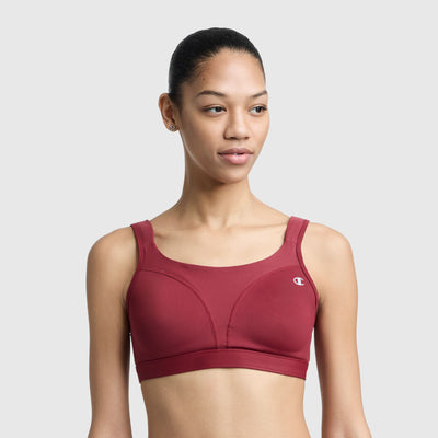A woman with tied-back hair wears the Champion Spot Comfort High-Impact Sports Bra, C Logo, in maroon. The Champion logo is on the left side. She stands against a light gray background, looking slightly to the side and smiling softly.