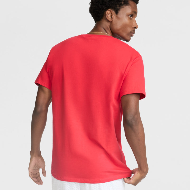 A man faces away from the camera, wearing a bright red Champion Classic T-Shirt with the C Logo and white shorts, set against a plain light backdrop. He slightly turns his head to the right, emphasizing its timeless men's style.