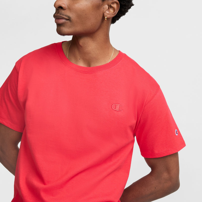A man wears a bright red Champion Classic T-Shirt, C Logo, with short sleeves and embroidered logos on the chest and sleeve, standing against a plain light background.