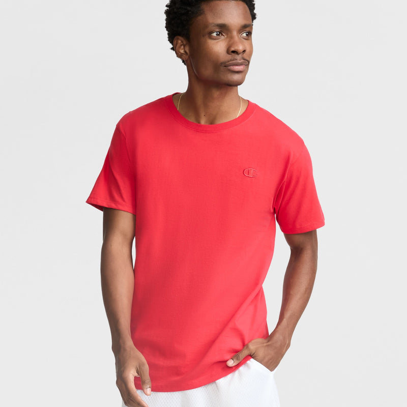 A man wearing the Champion Classic T-Shirt, C Logo in red with white shorts stands against a light gray background, looking slightly to his left with one hand in his pocket.