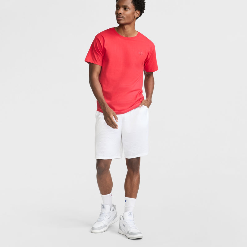 A man stands against a plain white background wearing a red Champion Classic T-Shirt with C Logo, white athletic shorts, socks, and sneakers. One hand is in his pocket as he looks to the side.