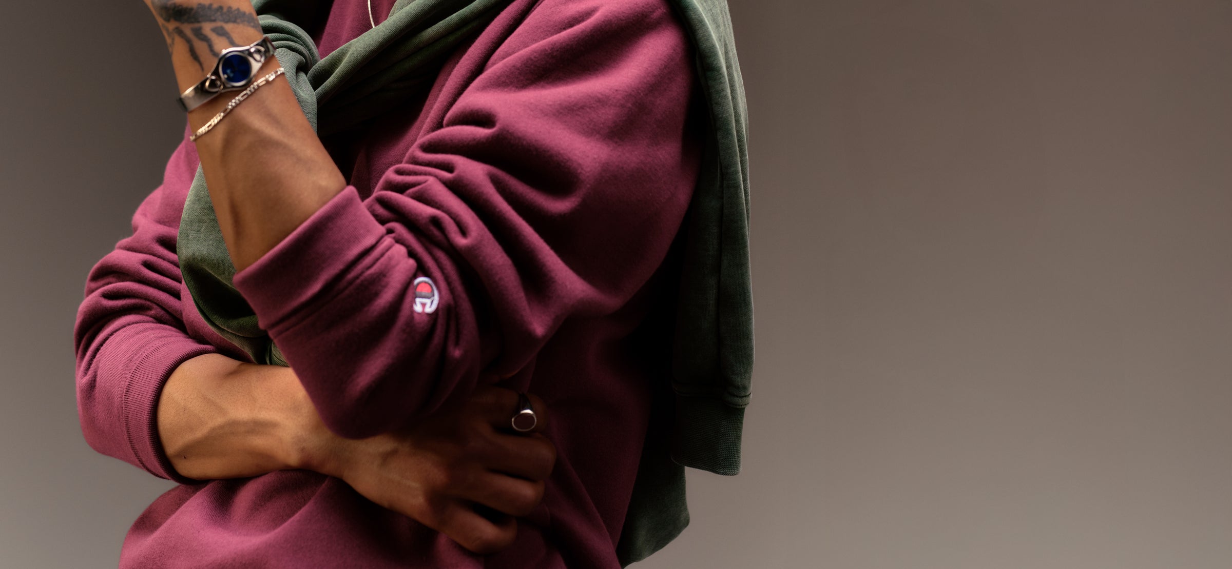 Close-up of a person with crossed arms wearing a maroon sweatshirt with an embroidered logo on the sleeve and a green sweatshirt draped over their shoulders, showcasing a bracelet, ring, and tattoos on their left arm.