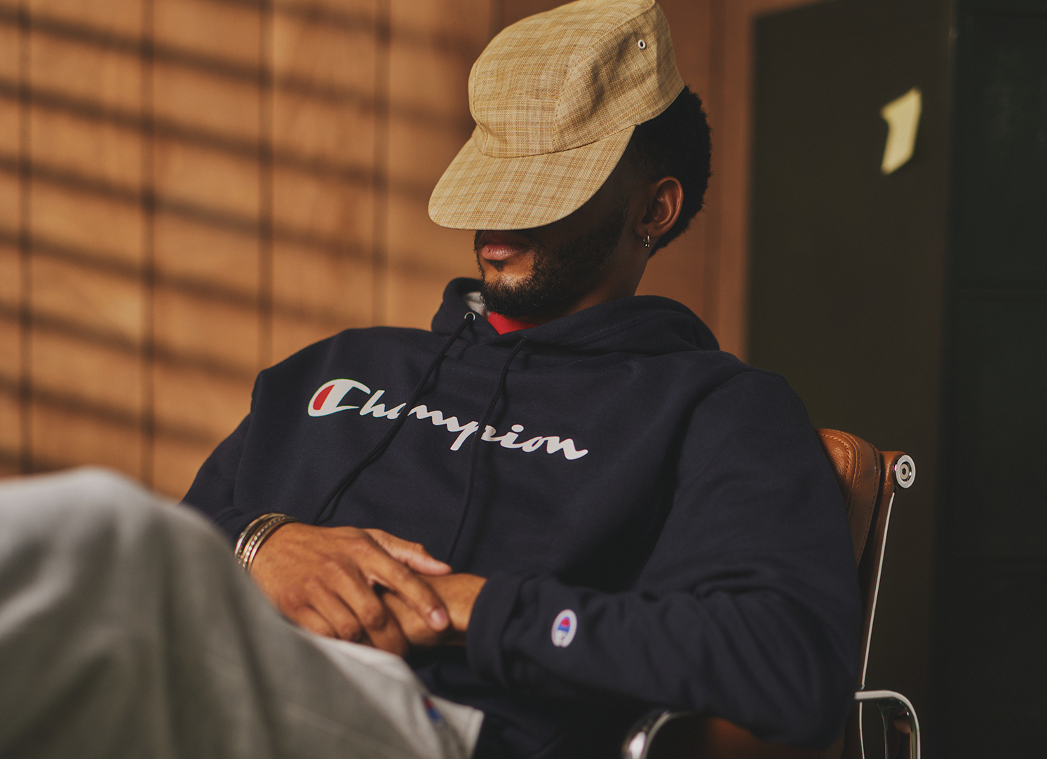 A person wearing a plaid cap and a navy Champion hoodie sits in a chair with their arms crossed and head tilted down, partially covering their face. Warm indoor lighting and shadows are visible in the background.