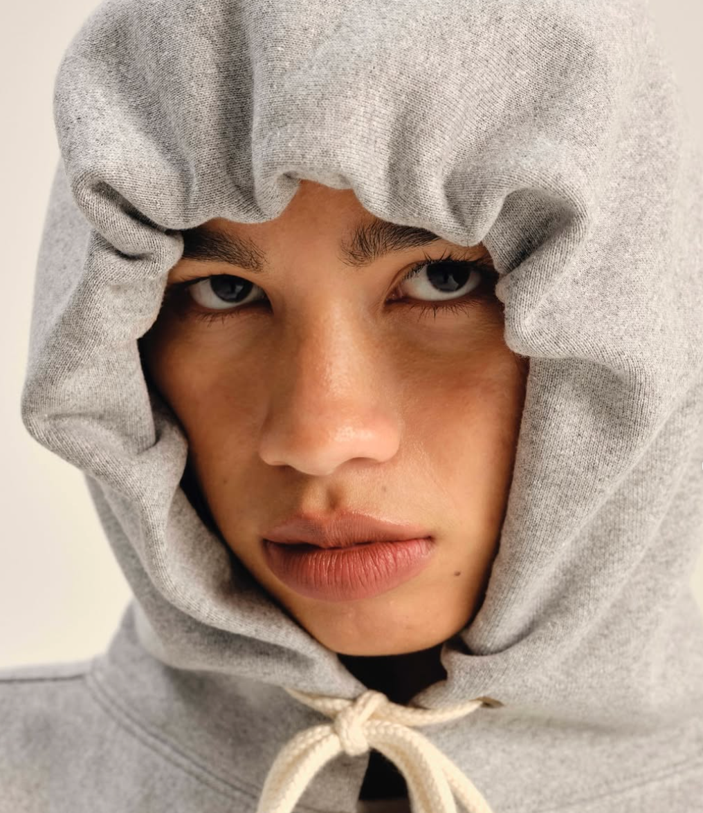 A person with a serious expression wears a gray hoodie pulled tightly around their face, framing only their eyes, nose, and mouth.