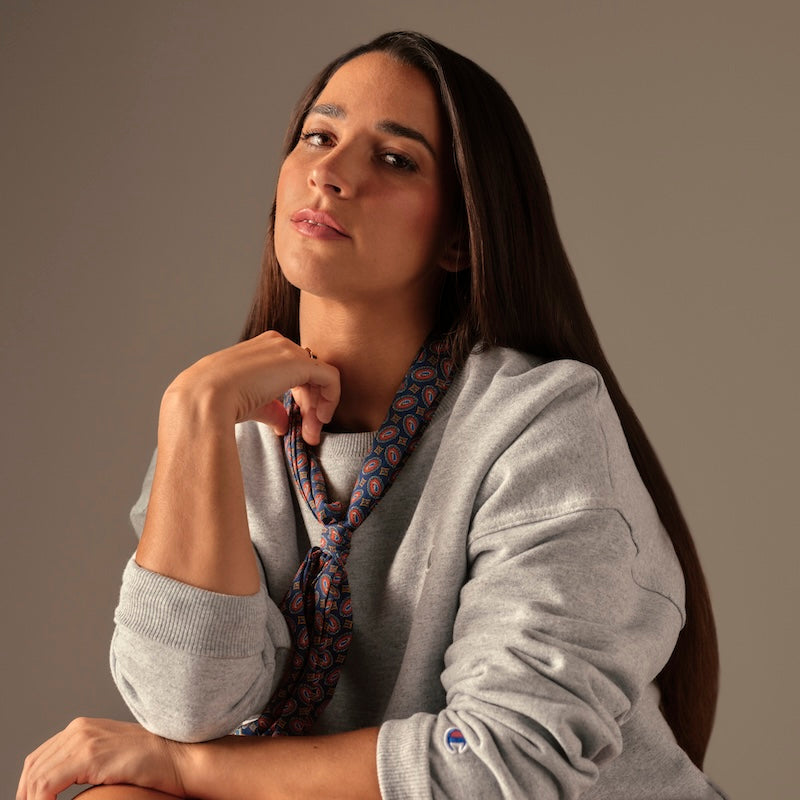 A woman with long straight brown hair wears a grey sweatshirt and a patterned scarf. She gazes confidently at the camera with her chin resting on her hand against a plain background.