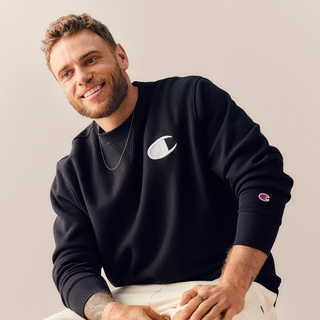 A smiling man with short curly hair and a beard wears a black Champion sweatshirt and light pants, sitting against a plain beige background. He has a gold chain necklace and is looking slightly to the side.