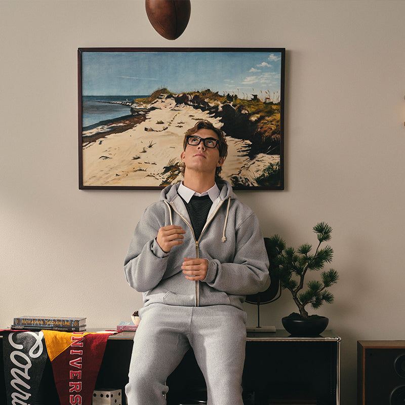 Person wearing a gray tracksuit standing in a room with a painting of a beach scene on the wall.