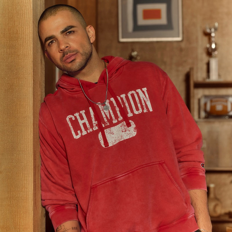 A man with a shaved head and trimmed beard leans against a wooden wall, wearing a faded red Champion hoodie and a necklace. Shelves with trophies and framed art are visible in the background.