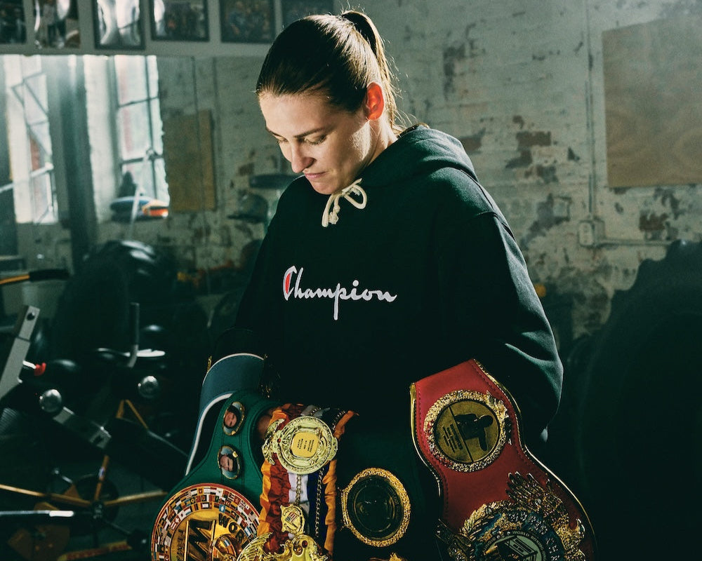 A person in a Champion hoodie stands indoors, looking down and holding several championship belts draped over their arms. The background shows gym equipment and a textured brick wall.