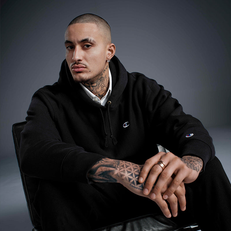 Man wearing a black hoodie with visible tattoos on his arms against a dark background