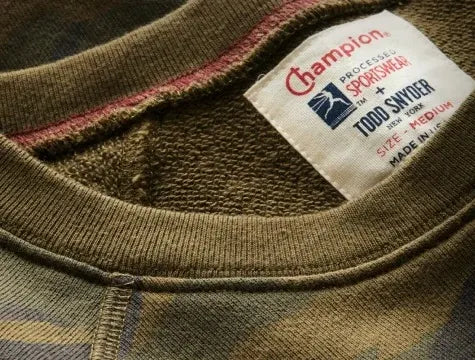 Close-up of a camouflage-patterned sweatshirt collar with a visible white tag showing Champion, Todd Snyder, and Medium size branding. The fabric appears worn and textured.