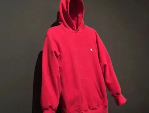 A red hooded sweatshirt with a small white Champion logo on the chest hangs against a plain dark wall. The hood is up and the sleeves are empty, making the hoodie appear unoccupied.