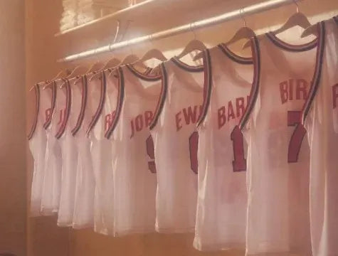 Several white basketball jerseys with red lettering and numbers hang on wooden hangers in a row inside a locker room. The jerseys display different names and numbers on the back.