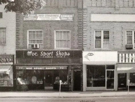 Black and white photo of a row of storefronts, including “Moe Sport Shops” with large front windows and a sign above the door, flanked by other shops on either side on a city street.