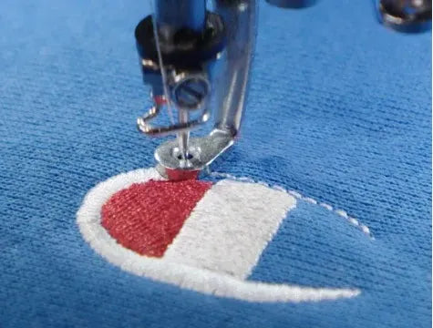 A sewing machine needle embroidering the red and white Champion logo on a blue fabric.