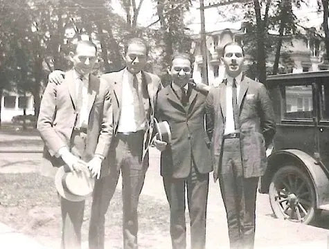 Four men in suits stand close together, smiling, outdoors near a vintage car and trees. Two men hold hats in their hands. Houses are visible in the background. The photo is black and white, giving a vintage feel.
