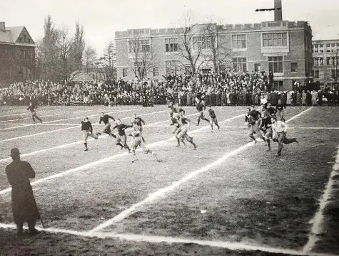 Black and white photo of a football game on a grassy field, with players running and tackling. A large crowd stands in the background near brick buildings, and a person in a coat watches from the sideline.