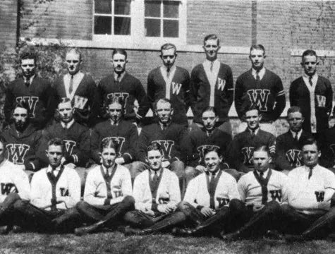 A black-and-white photo of a vintage sports team. Three rows of young men pose outdoors, most wearing sweaters with large W letters and neckties, in front of a brick building with windows.