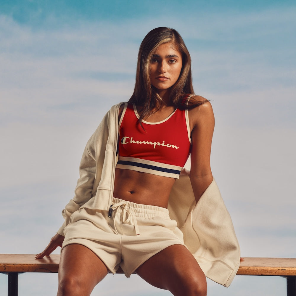 A woman sits on a bench against a blue sky backdrop, wearing a red Champion sports bra, cream-colored shorts, and a matching cream jacket draped off her shoulders. She looks confidently at the camera.