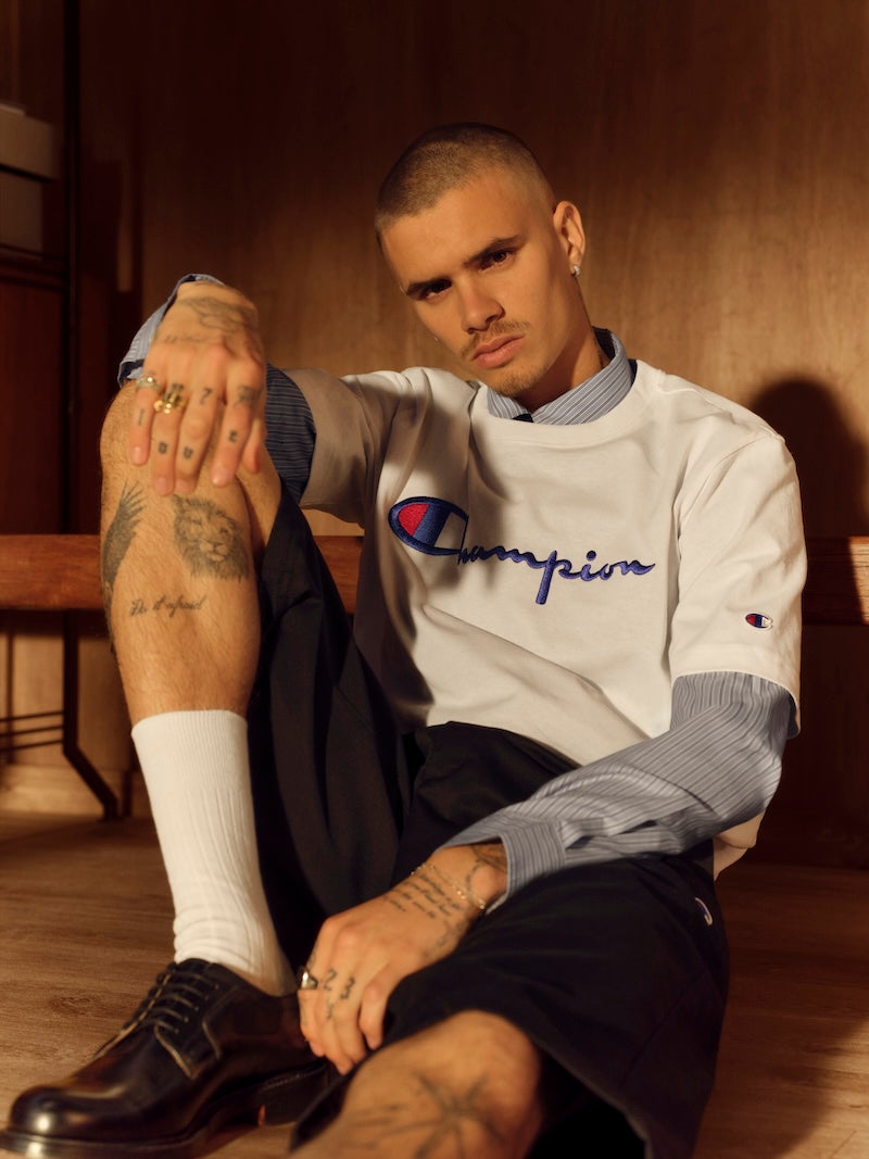 A man with a buzz cut, tattoos on his hands and legs, and multiple rings sits on the floor. He wears a white Champion t-shirt over a striped shirt, black shorts, white socks, and black shoes in a warmly lit room.