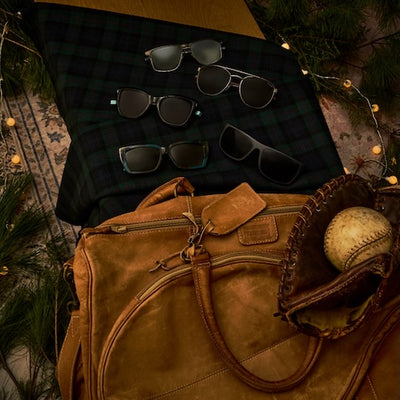 Brown leather bag with sunglasses and a baseball on a checkered blanket.