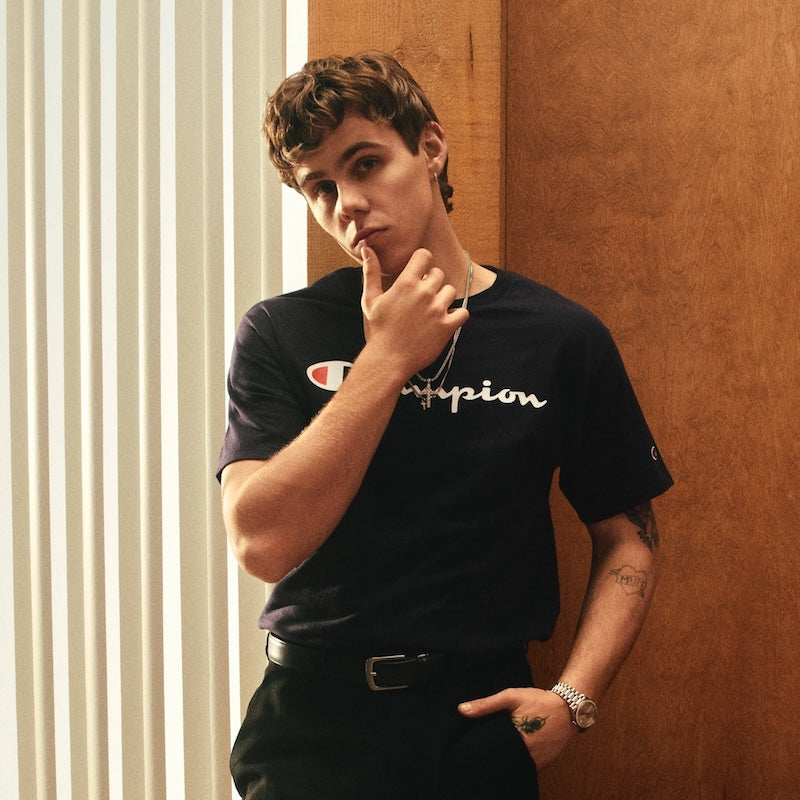 A young man with short brown hair wearing a navy blue Champion t-shirt stands indoors, touching his lips thoughtfully with one hand. He has tattoos on his left arm, wears a watch, and stands in front of wooden and vertical blind backgrounds.